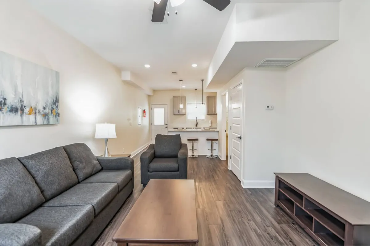 The image showcases a stylish, modern living area featuring a comfortable sofa, a sleek coffee table, inviting kitchen bar seating, and striking wall art. Soft lighting from a nearby lamp creates a welcoming ambiance, perfect for relaxing or entertaining guests.