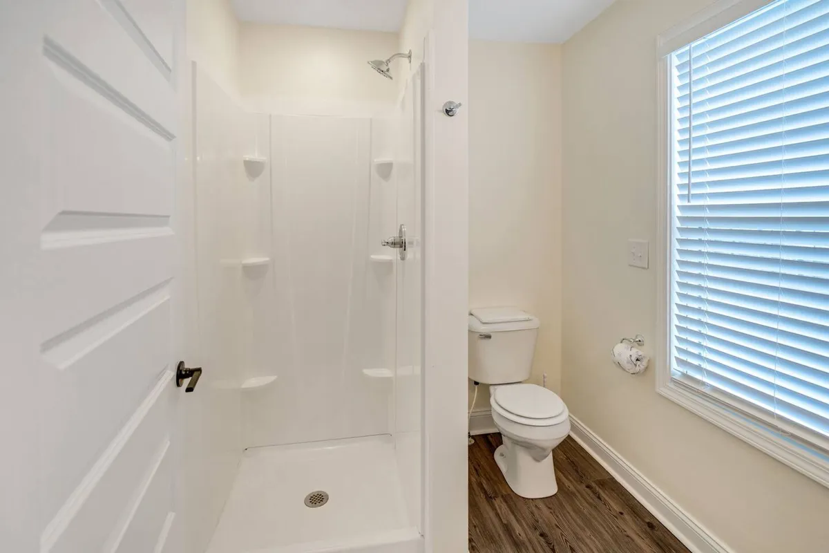 A fresh, inviting bathroom featuring a spacious shower area, toilet, and a window with blinds that allow natural light to filter in. The mood is clean and refreshing, suggesting both relaxation and utility.