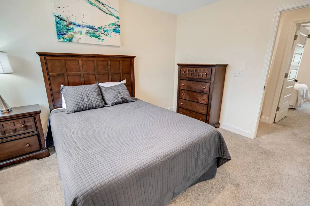 A cozy bedroom featuring a bed with a gray quilt, complemented by a wooden nightstand and dresser. Abstract wall art adorns the walls, and the ambiance is restful and inviting—ideal for relaxation or unwinding with a book.