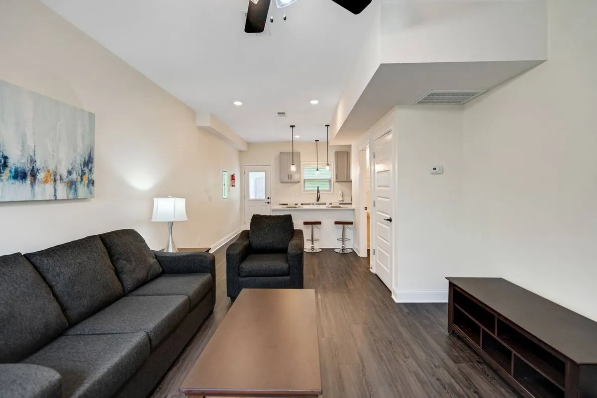 A spacious interior featuring a sofa, armchair, coffee table, visible kitchen area, and modern artwork. The layout encourages both relaxation and entertaining, and the space is bathed in natural light.