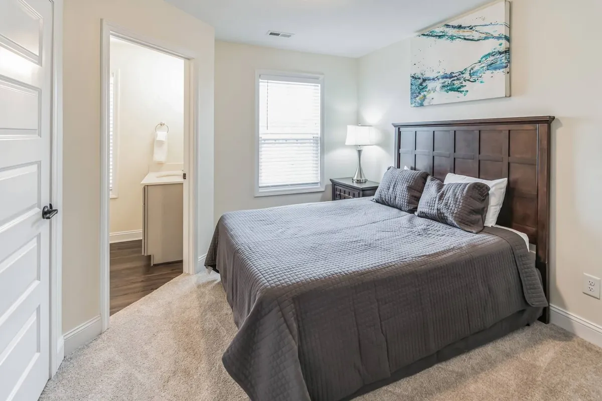 A serene bedroom scene with a dark wood headboard, elegant artwork above the bed, and a softly lit lamp on the bedside table. Sunlight streams in, highlighting the cozy comfort that's perfect for relaxing after a day out.