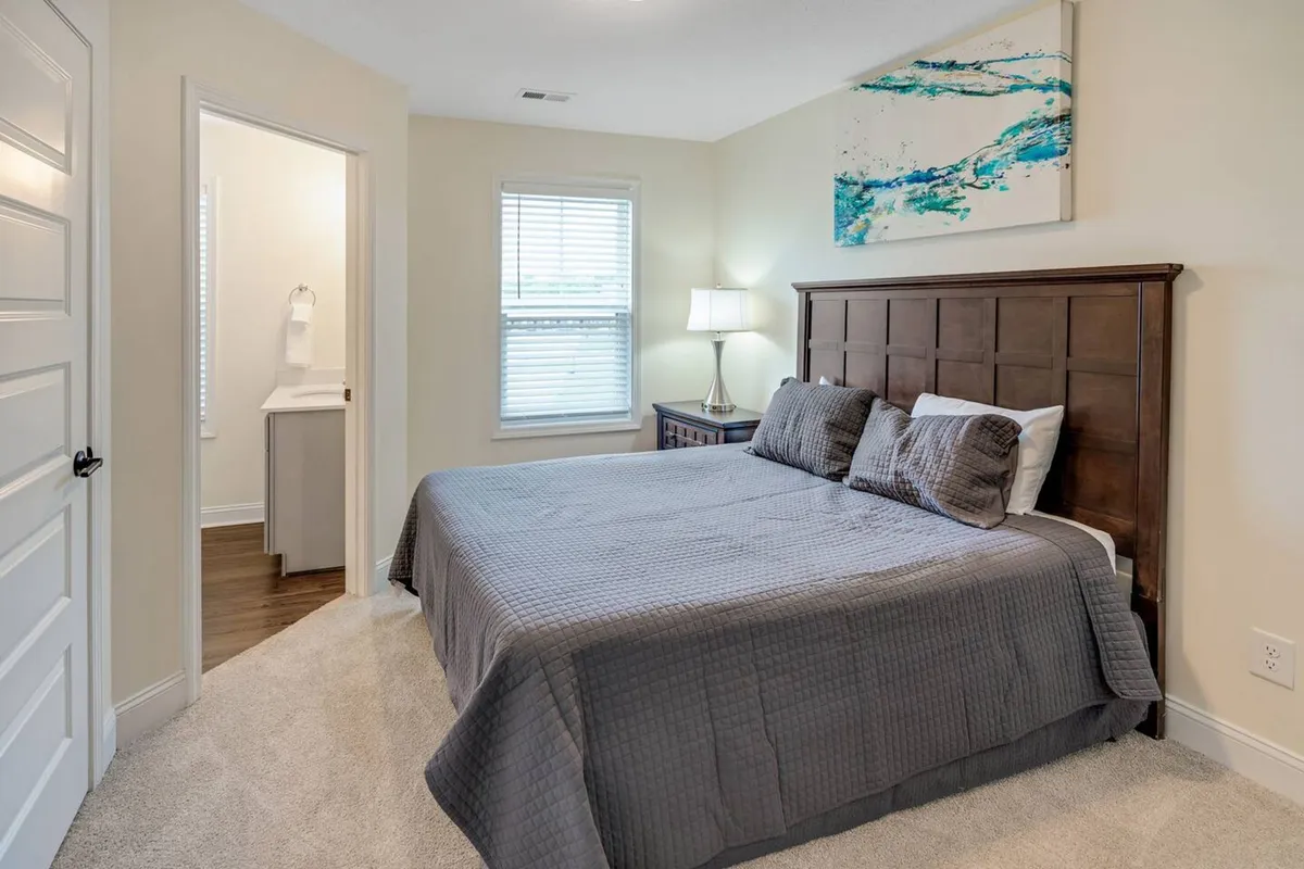 A serene bedroom with a gray-quilted bed, wooden headboard, softly glowing side lamp, and abstract wall art. The doorway leads directly to a bathroom, highlighting privacy and ease of access for guests.