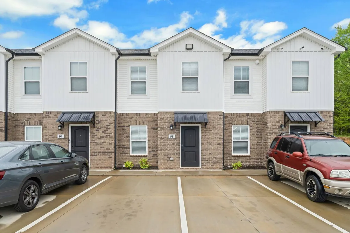 The image features the welcoming exterior of a townhouse with well-maintained landscaping and a visible parking area. The inviting porch and front facade suggest a friendly neighborhood, while the open outdoor space hints at opportunities for morning coffee or casual gatherings.