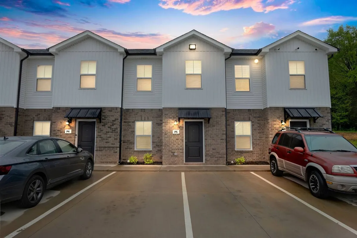 This image depicts the exterior front entrance of the property, featuring a welcoming black door, evenly spaced windows, and convenient parking spaces. The scene alludes to neighborhood gatherings and residents enjoying the outdoors, with touches like sunset views from the porch and friendly chats with neighbors.